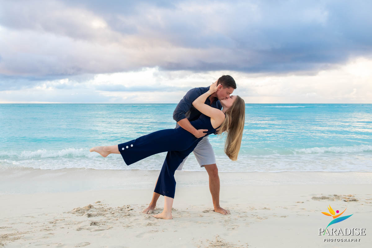Man dips partner for kiss under cloudy skies along turquoise coastline